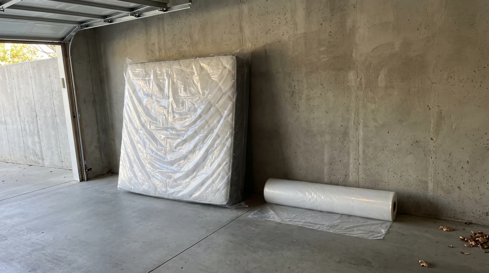 A plastic-wrapped mattress leaning against a bare garage wall with a roll of clear sheeting on the concrete floor, ready for furniture removal in Meridian, Idaho