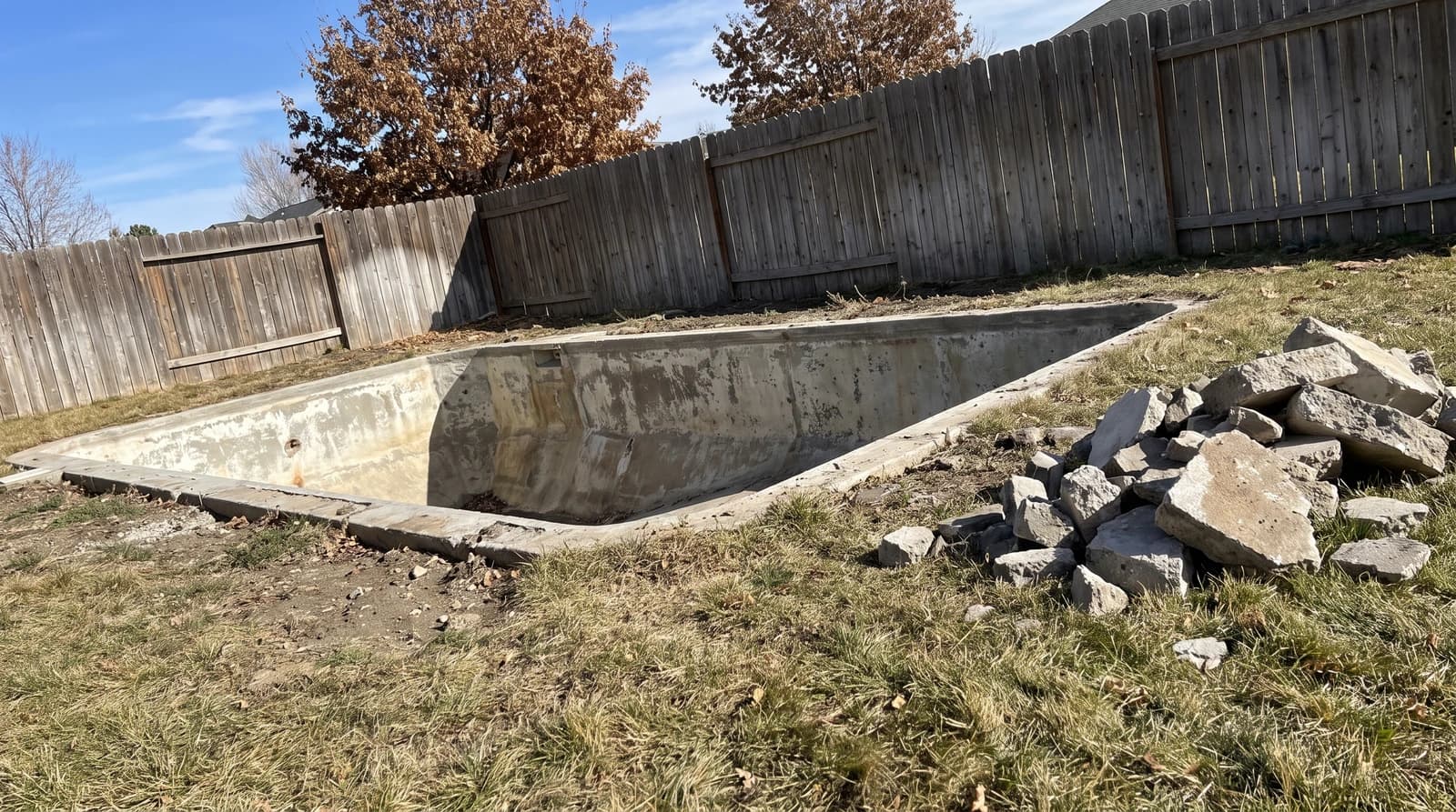 Empty concrete pool shell with broken concrete piled on the grass beside a wooden privacy fence during a pool demolition in Boise, Idaho