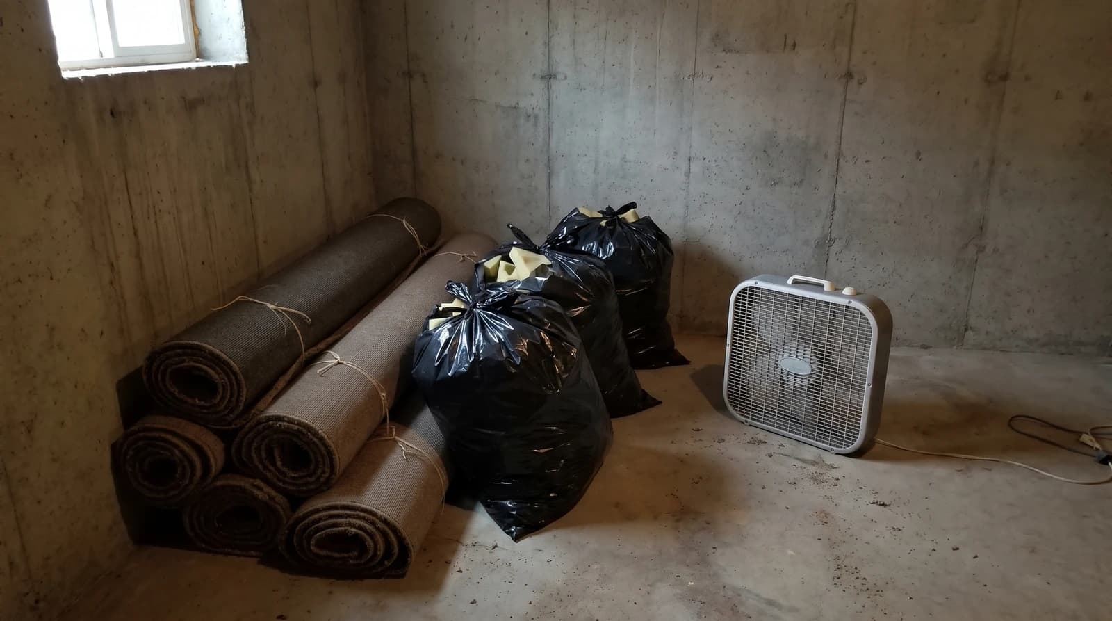 Rolled wet carpet strips and bagged carpet pad on a bare concrete basement floor after a water leak in Boise, Idaho