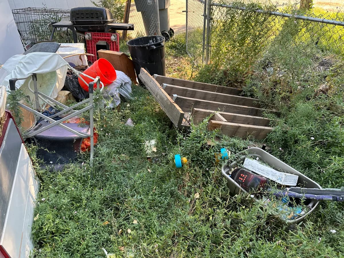 Overgrown yard filled with an old grill folding chairs wooden crates buckets and debris at a property needing yard cleanout in Star Idaho