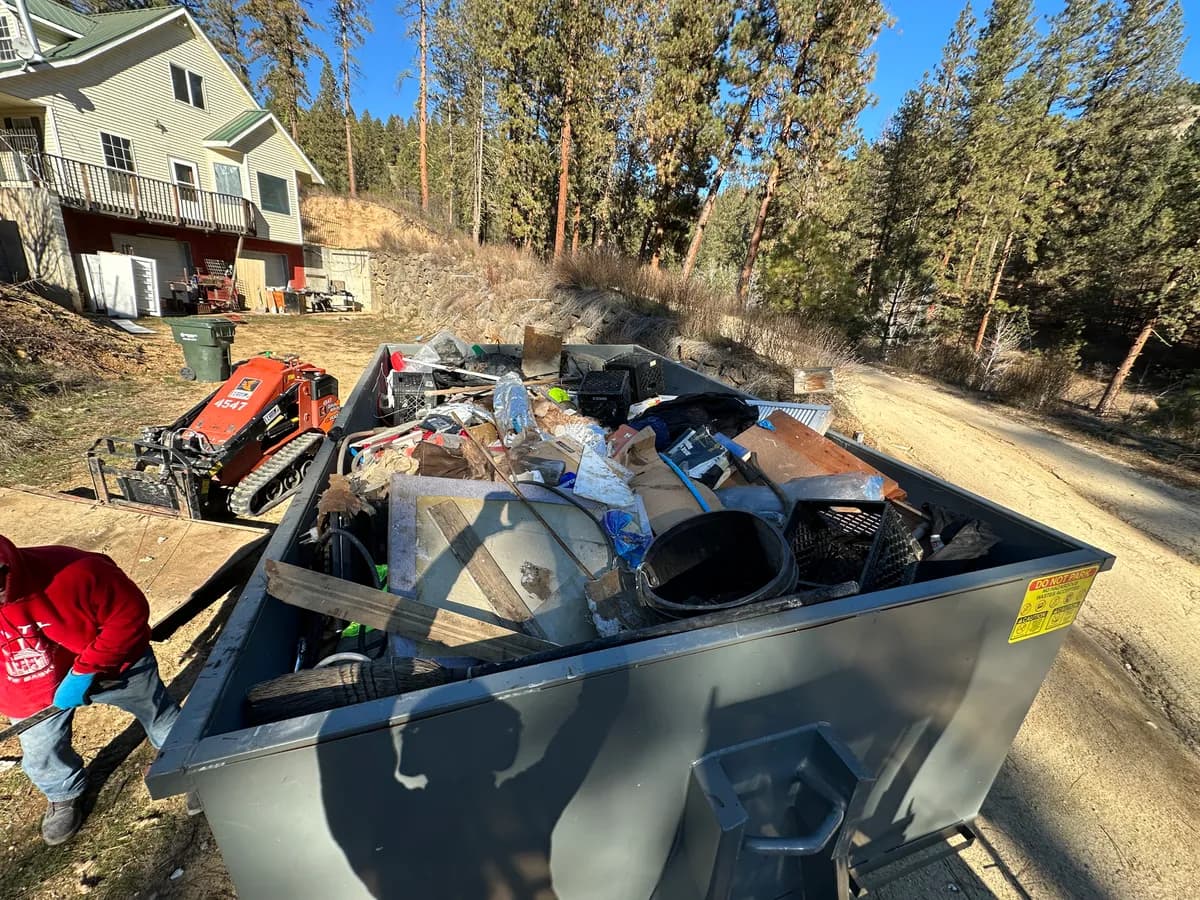 Top Shelf crew with a full dumpster and mini track loader clearing property debris from a wooded hillside lot in Middleton Idaho