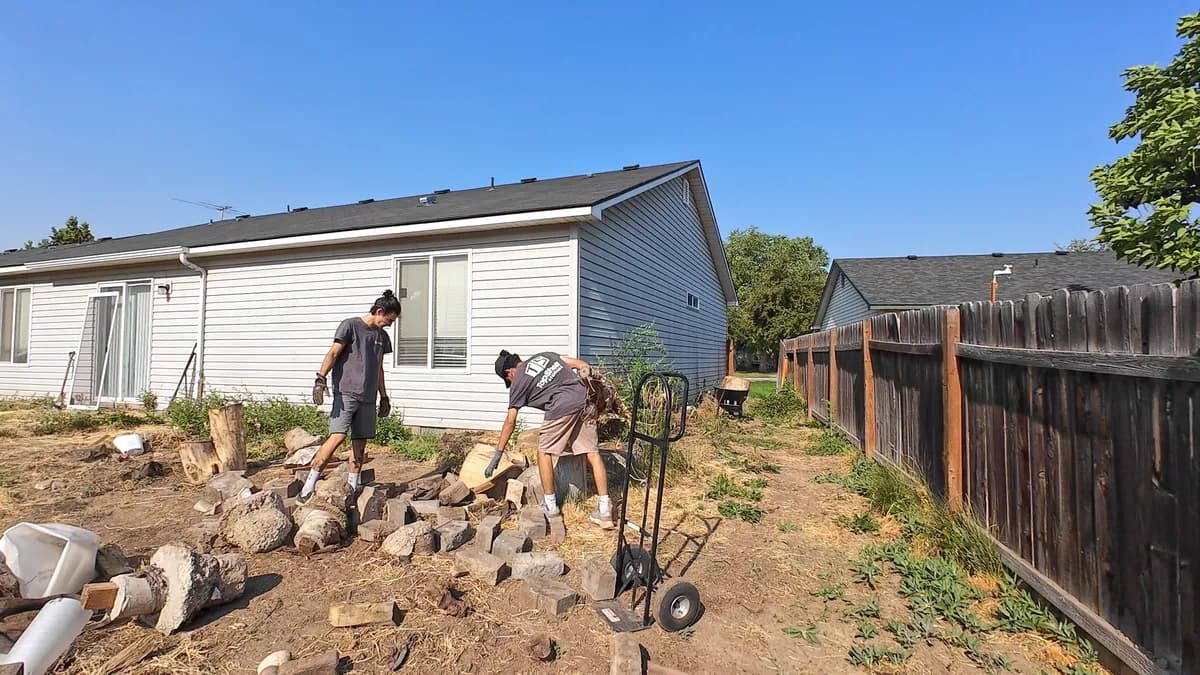 Two Top Shelf crew members clearing concrete chunks and wood debris from a residential backyard with a hand truck in Kuna Idaho