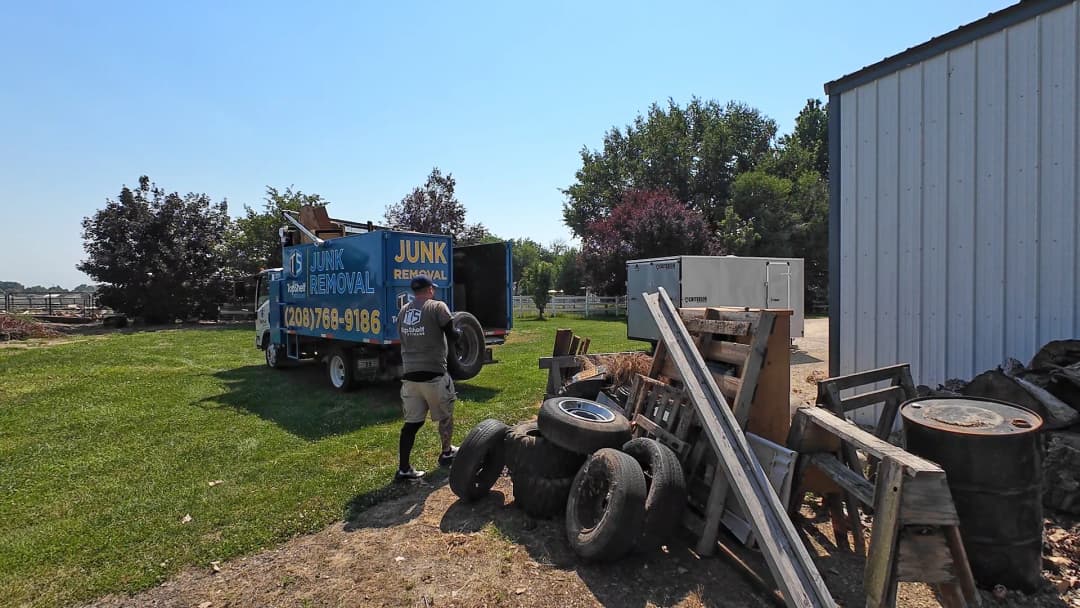 Top Shelf crew member carrying a tire past oil drums and scrap debris to the branded truck at a rural property in Caldwell Idaho