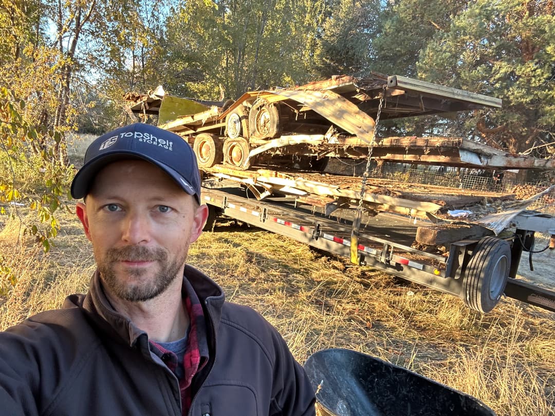Top Shelf crew member in branded hat standing next to a flatbed trailer loaded with heavy machinery and demolition debris in Eagle Idaho