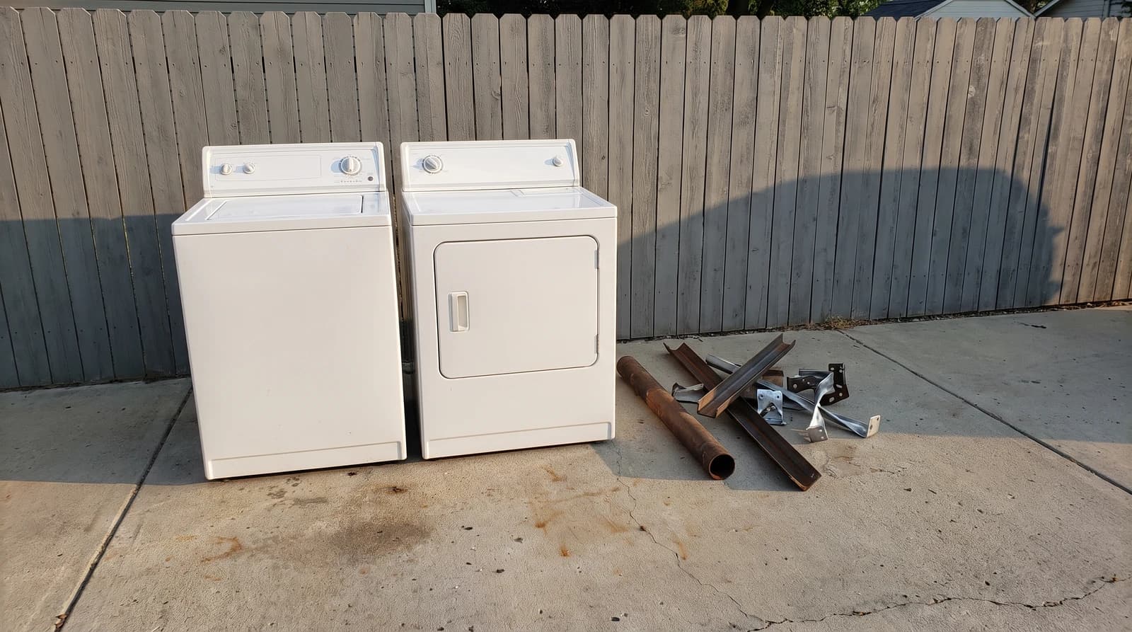 An old washer and dryer set on a concrete driveway beside a stack of scrap metal, ready for appliance removal at a home in Nampa, Idaho.