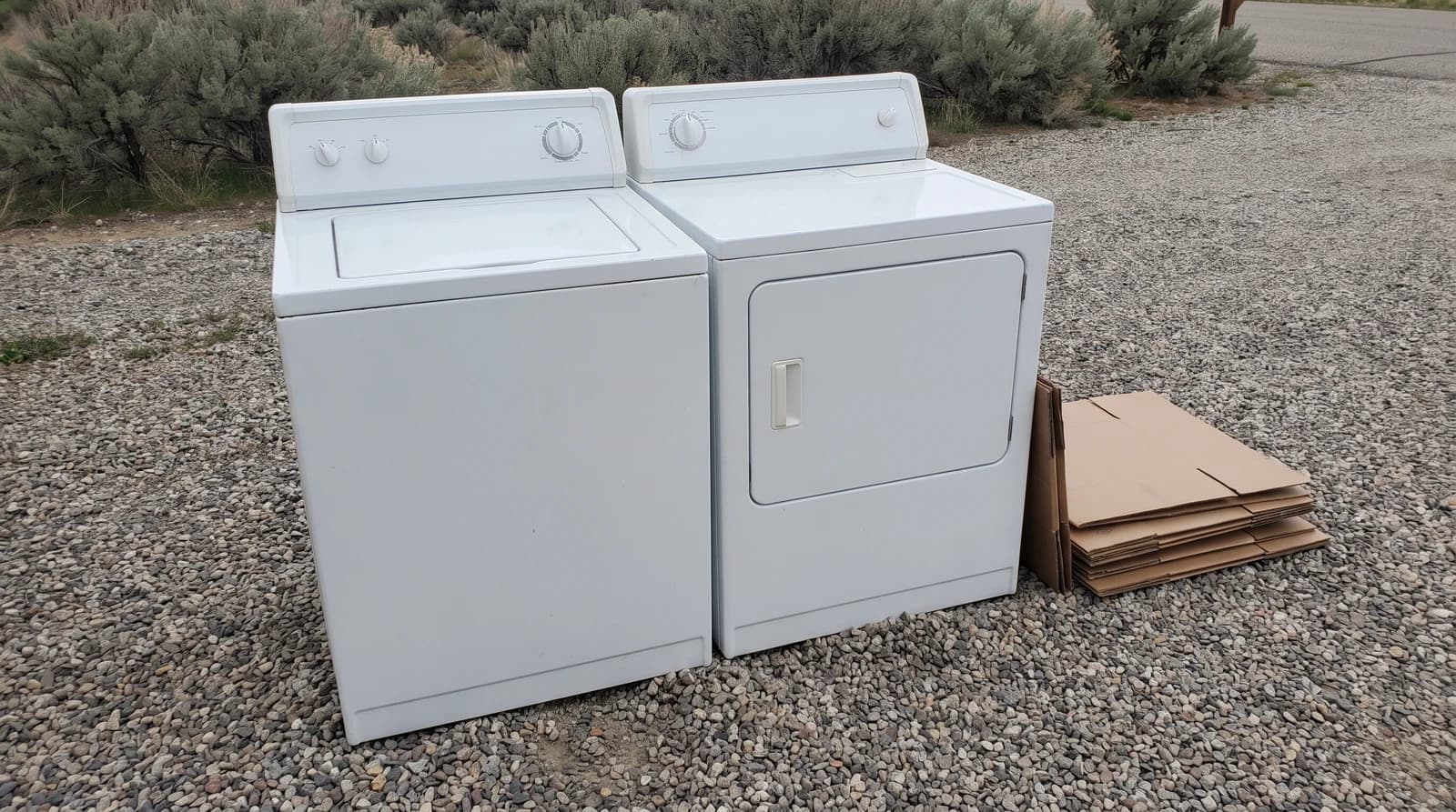 An old washer and dryer set on a gravel driveway beside stacked cardboard, waiting for appliance removal at a home in Star, Idaho.