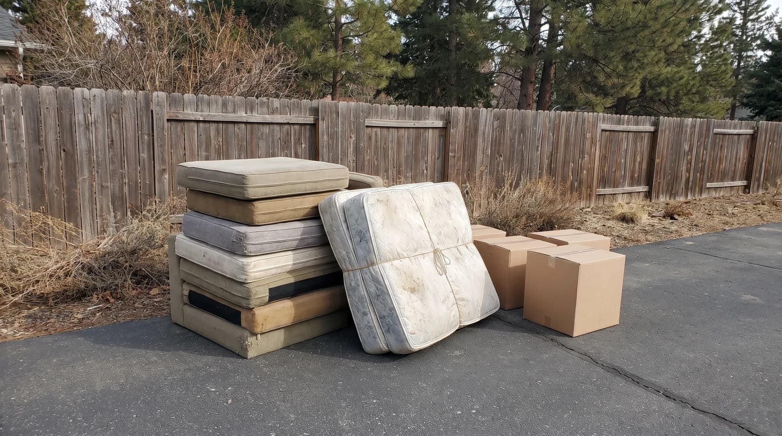 Old couch cushions, a mattress and cardboard boxes stacked on pavement beside a wooden fence at an apartment complex in Boise, Idaho, ready for bulk trash removal
