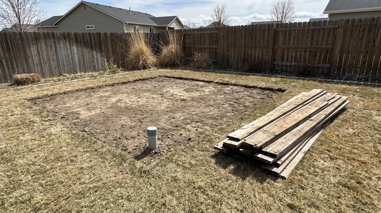 Bare dirt footprint and capped conduit stub left after a backyard shed demolition in Kuna, Idaho, with stacked wood siding boards on the grass