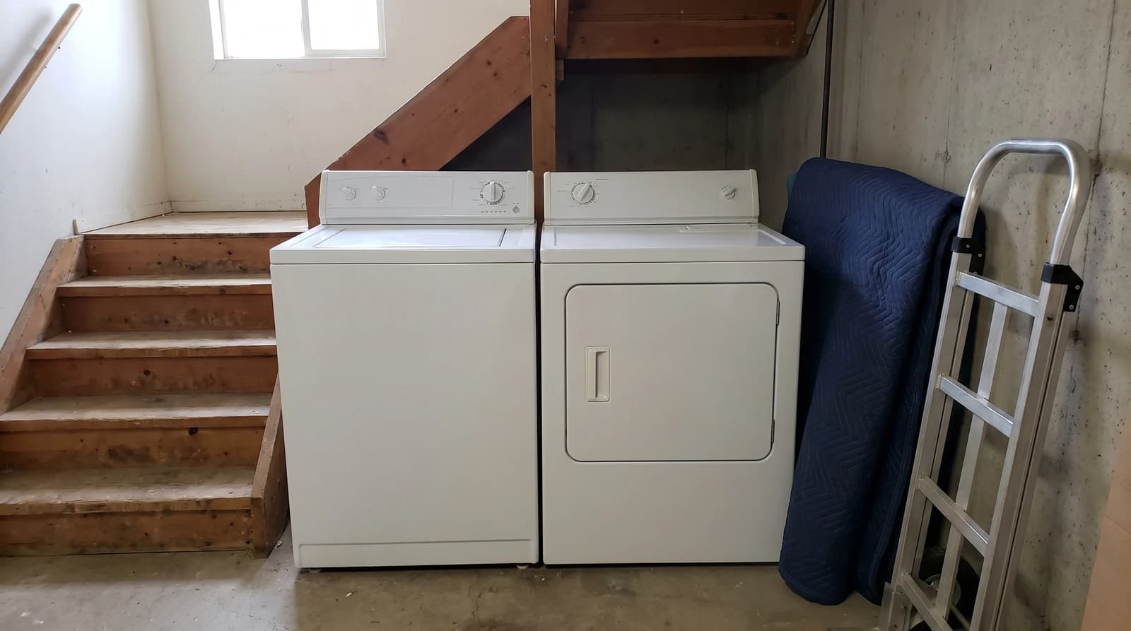 Old white washer and dryer at the bottom of a narrow basement staircase in a Boise, Idaho home, with a moving blanket and hand dolly ready for appliance removal