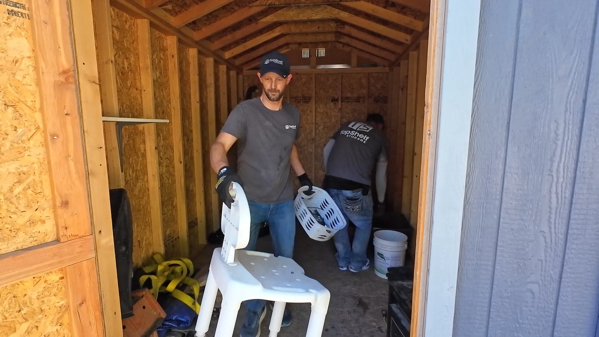 Two Top Shelf crew members in branded shirts clearing out a storage shed carrying chairs and household items in Garden City Idaho