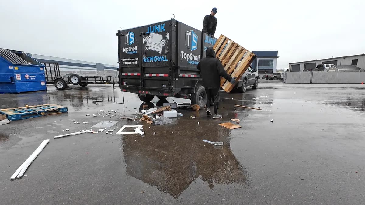 Two Top Shelf crew members loading wooden pallets into the branded junk removal trailer at a commercial lot in Emmett Idaho
