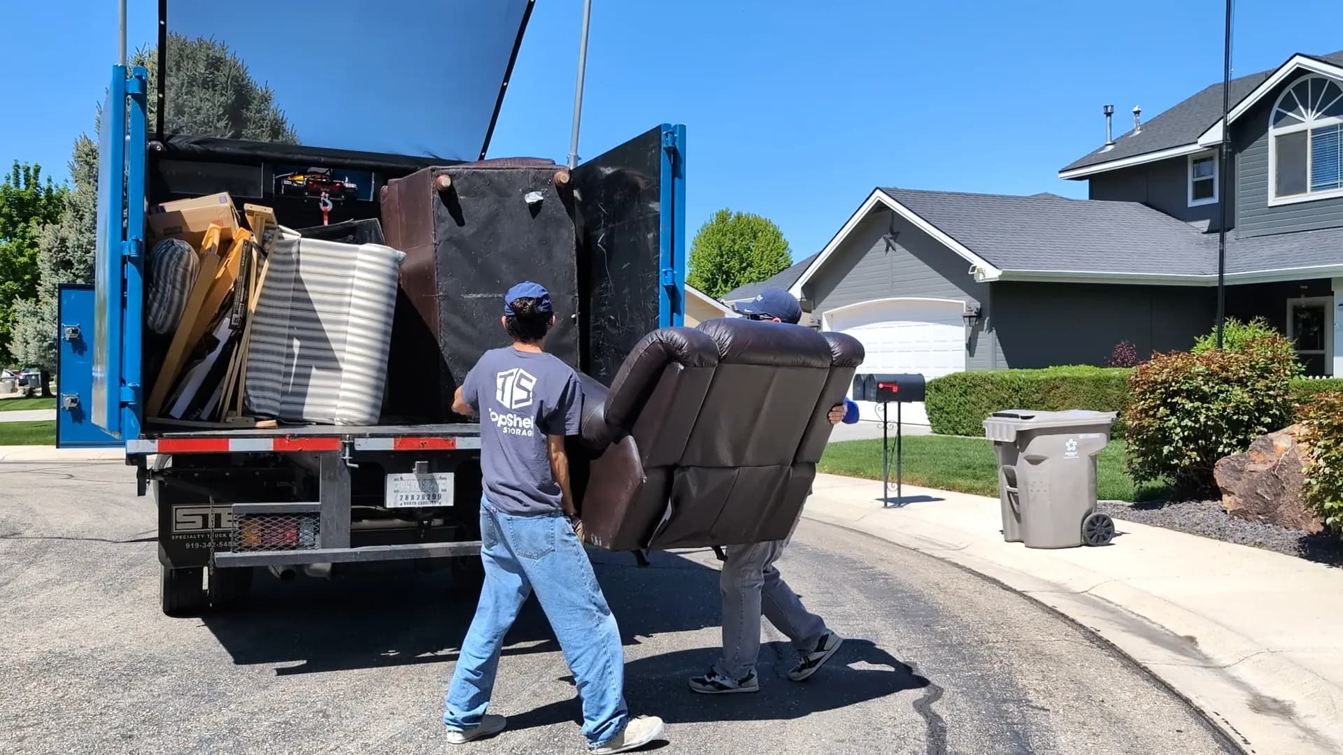 Two Top Shelf crew members in branded shirts lifting a brown leather recliner into the junk removal truck in a subdivision in Eagle Idaho