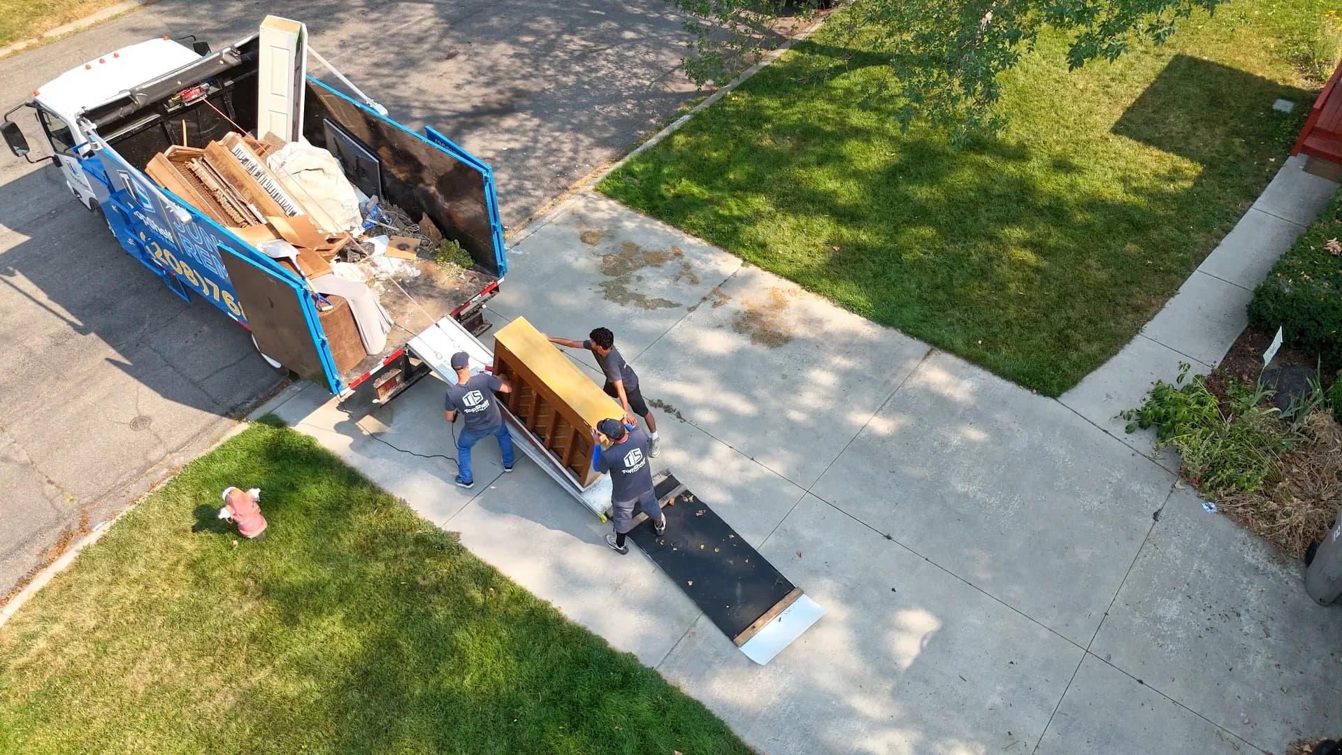 Aerial view of three Top Shelf crew members in branded shirts loading an upright piano up the ramp into the junk removal truck at a residential property in Star Idaho