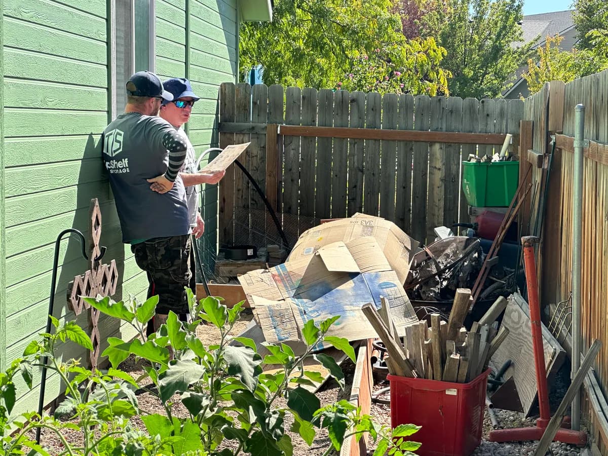 Two Top Shelf crew members assessing a backyard clutter pile with cardboard and miscellaneous junk in Garden City Idaho